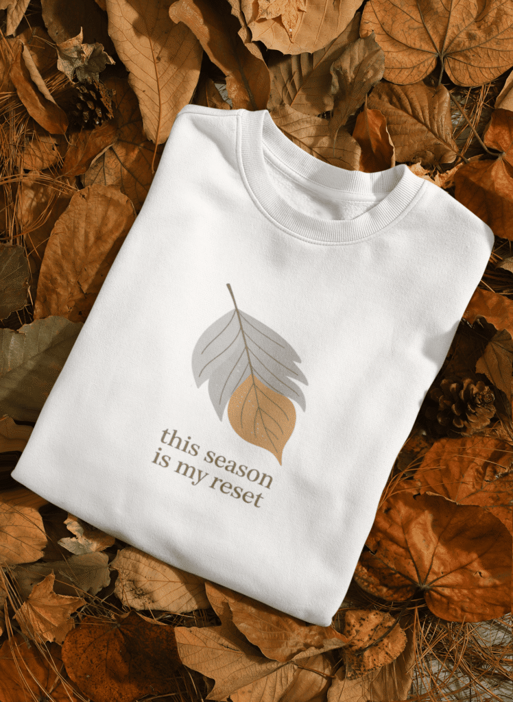 White sweatshirt with a minimalist autumn leaf design and the text ‘This season is my reset,’ folded on a bed of fall leaves, cozy autumn apparel mockup.