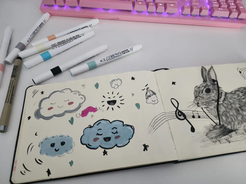 Inside My Sketchbook: A Real Look Behind the Art 2 Open sketchbook with cute cloud doodles, pastel hearts, and a detailed bunny drawing, surrounded by scattered markers and a pink keyboard in the background.