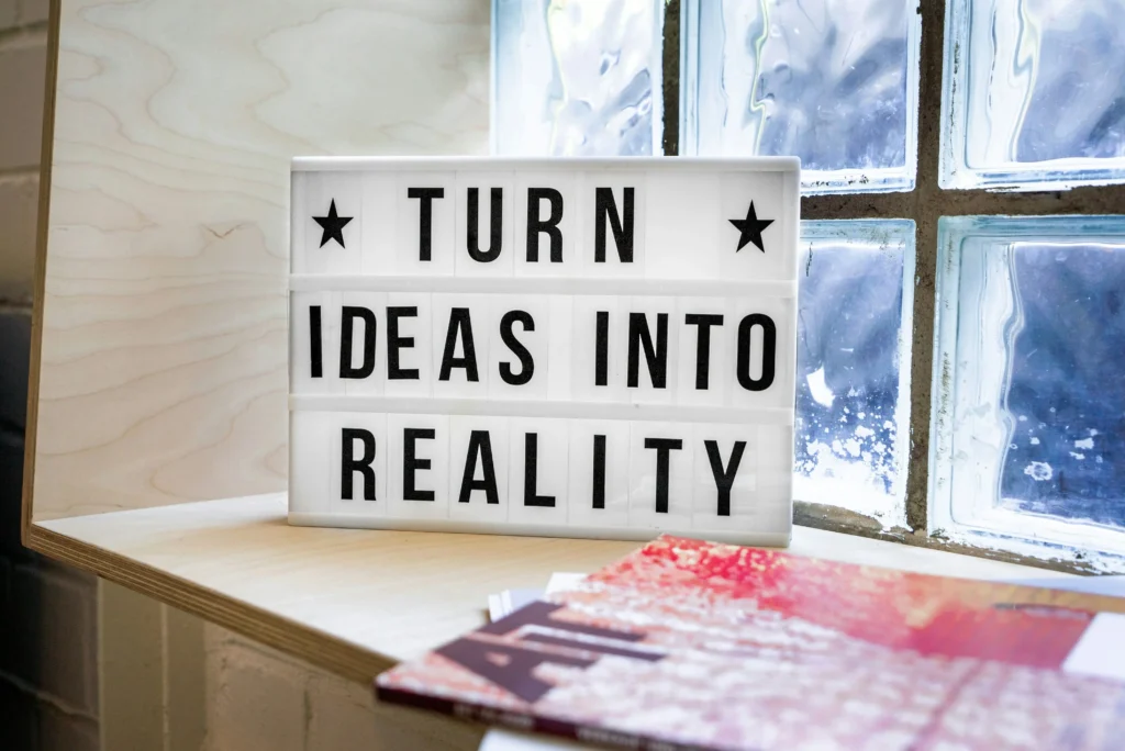 Why It’s Okay to Start Small (And Stay Consistent) 3 Lightbox sign with bold black text that says “Turn Ideas Into Reality,” placed on a wooden shelf by a frosted window.
