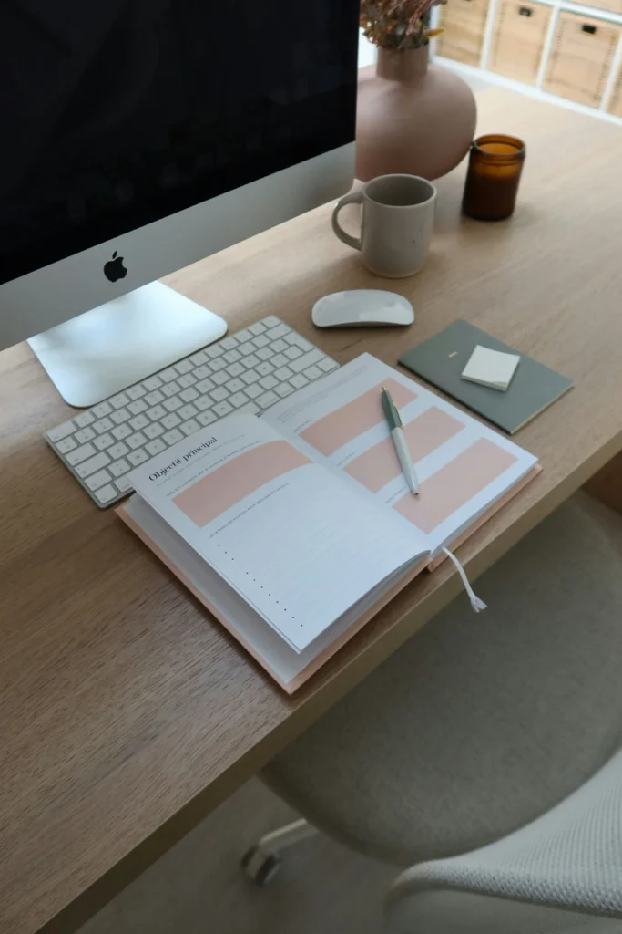 Simple morning routine setup with a pink planner, cup of coffee, and iMac on a wooden desk, evoking calm and productivity.