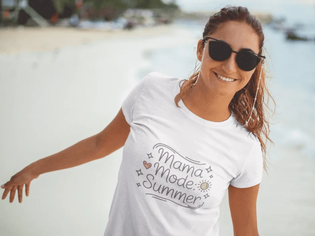 Smiling woman at the beach wearing white t-shirt with Mama Mode: Summer design and black sunglasses – summer vibe mockup