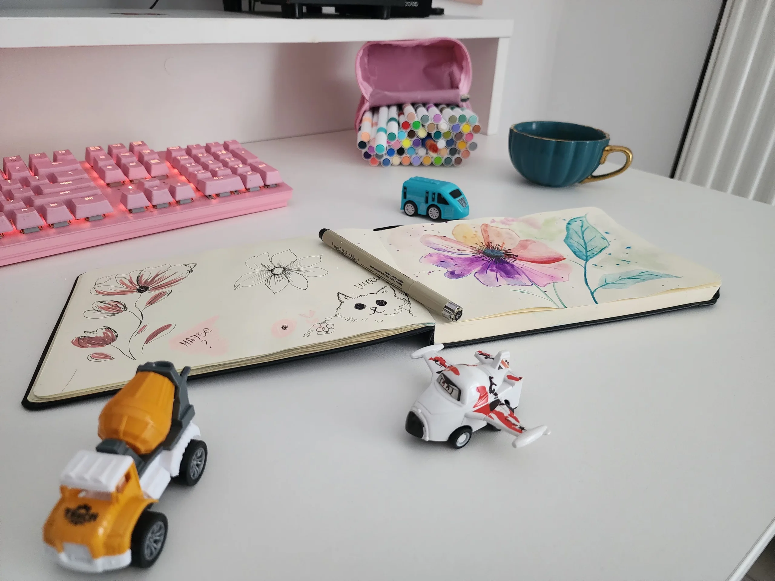 Open sketchbook with watercolor flowers on a mom's desk, surrounded by toddler toys, pink keyboard, and a cup of coffee – capturing everyday creativity in a cozy workspace.