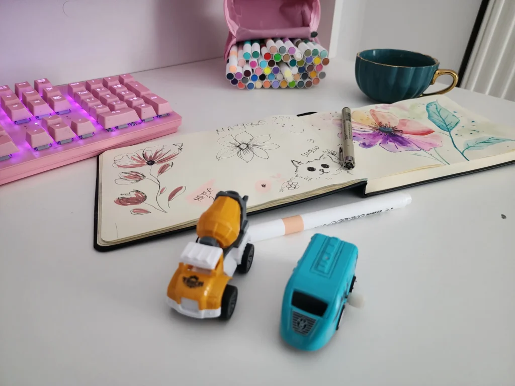 Inside My Sketchbook: A Real Look Behind the Art 1 Open sketchbook with messy flower doodles and the word “maybe?”, surrounded by markers, toy cars, and a pink keyboard — a real look inside my sketchbook as a creative mom.