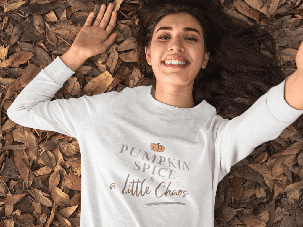 Smiling woman lying on autumn leaves wearing a white sweatshirt with the text ‘Pumpkin Spice & a Little Chaos,’ cozy fall apparel.